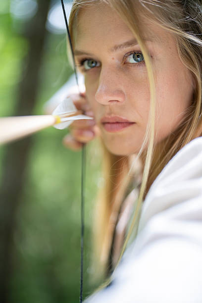 Young Caucasian female archer outdoors in medieval attire, holding a bow with focused determination as she aims carefully among the trees.