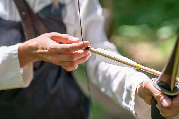 Young Caucasian woman in a medieval setting carefully preparing to shoot an arrow with a bow, focusing intently as sunlight filters through the surrounding foliage.