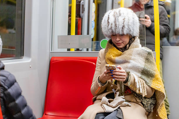 Pretty female passenger in winter attire, including a fur hat and coat, scrolling through her phone on the subway.