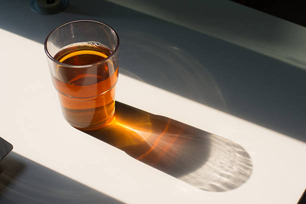 A mesmerizing close-up of sunlight passing through a glass of tea, creating a sharp, golden caustic light pattern and refraction on a white desk surface