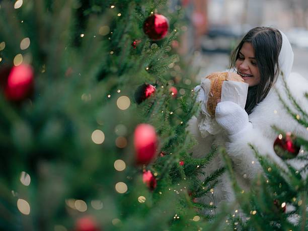 Close up portrait cheerful young woman in white winter coat ready to bite baked roll, framed by festive Christmas tree branches.