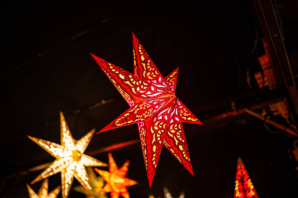 A close-up of a glowing red paper star lantern featuring detailed patterns, displayed at a Christmas market. Warm lighting enhances the festive seasonal atmosphere.