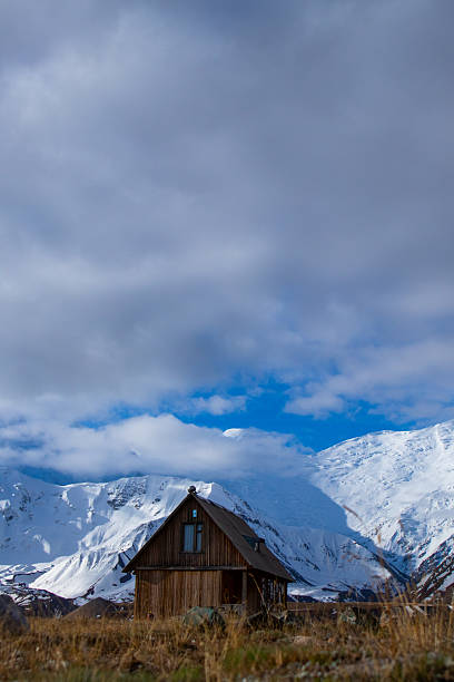 Rustic wooden cabin situated in a vast landscape beneath towering snow-covered mountain peaks under a cloudy sky