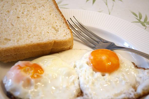 Two homemade fried eggs served on a white plate with bread slices in natural condition, photographed in a close-up style for simple homemade breakfast concepts.