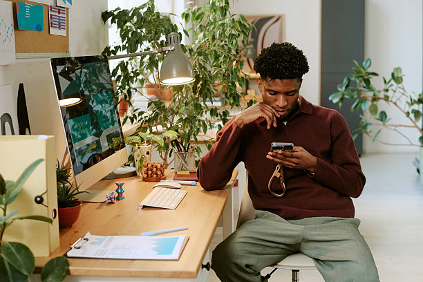 istock Young Adult Black Man Using Smartphone While Sitting at Modern Office Desk 2251350597