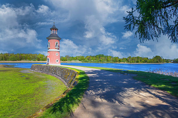 Dramatic shot of the Moritzburg Lighthouse by the pond. Brick-patterned facade under a cloudy blue sky. Path and saturated greenery in the foreground.