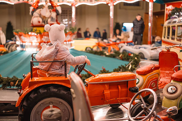 A bundled-up child enjoys a ride on a vintage orange tractor at an illuminated holiday carousel. Twinkling lights and decorated surroundings create a joyful and nostalgic winter fair atmosphere.