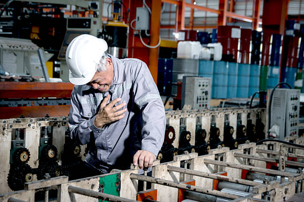 Asian senior foreman with safety helmet experiencing a heart attack while inspecting machinery in manufacturing factory. Elderly worker has cardio problems chest pain during working in industrial site