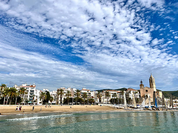 View of Passeig de la Ribera in Sitges with turquoise Mediterranean, sandy beach, white coastal buildings and sailboats under a blue summer sky.Sitges is a village about 35 kms. southwest of Barcelona, in Catalonia, Spain, renowned worldwide for its film festival, Carnival. Located between the Garraf Massif and the Mediterranean Sea, it is known for its beaches, nightspots, and historical sites. Sitges has been referred to as the Saint-Tropez of Spain, with property prices approaching those of the most expensive European cities.