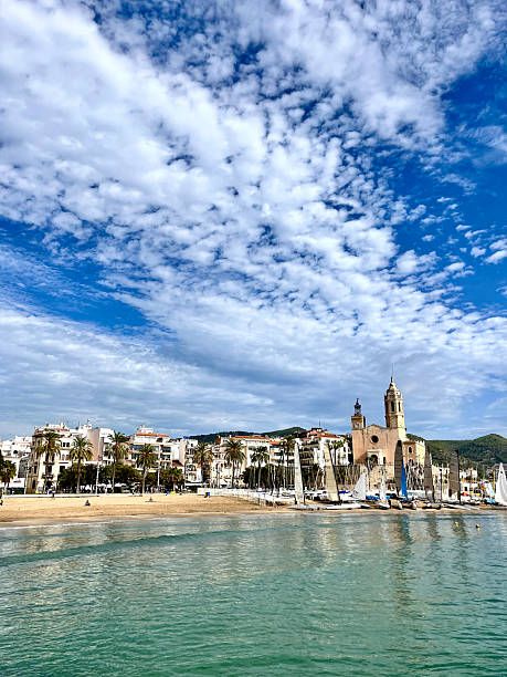 View of Passeig de la Ribera in Sitges with turquoise Mediterranean, sandy beach, white coastal buildings and sailboats under a blue summer sky.Sitges is a village about 35 kms. southwest of Barcelona, in Catalonia, Spain, renowned worldwide for its film festival, Carnival. Located between the Garraf Massif and the Mediterranean Sea, it is known for its beaches, nightspots, and historical sites. Sitges has been referred to as the Saint-Tropez of Spain, with property prices approaching those of the most expensive European cities.