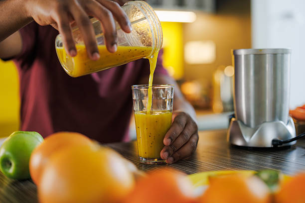 Close up of a man pouring fresh orange juice into a glass in a bright kitchen with fruits on the counter and a blender nearby, natural light illuminating the scene