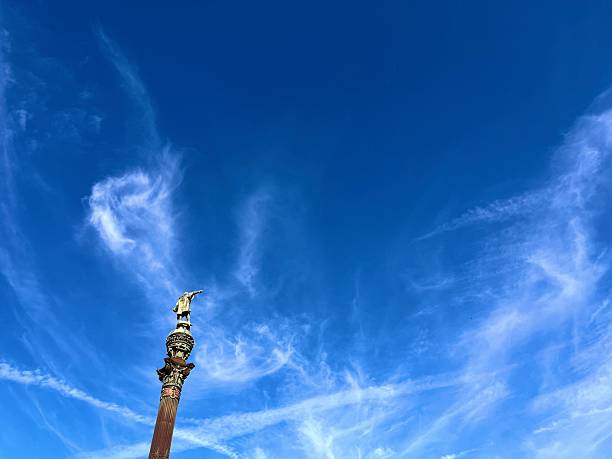 Statue of Christopher Columbus standing atop the Columbus Monument, reaching towards the sea under a vibrant blue sky with wispy clouds, symbolizing exploration, travel, and historical landmarks