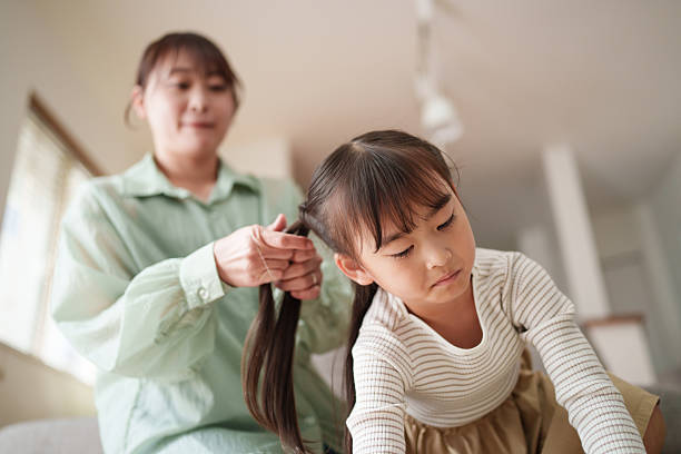 Mother carefully braids her young daughter’s hair in a cozy living room while the child sits calmly. The scene represents everyday family routines, parental care, tenderness, and quiet bonding moments at home. Ideal for concepts such as parenting, childhood, trust, domestic life, and emotional connection.