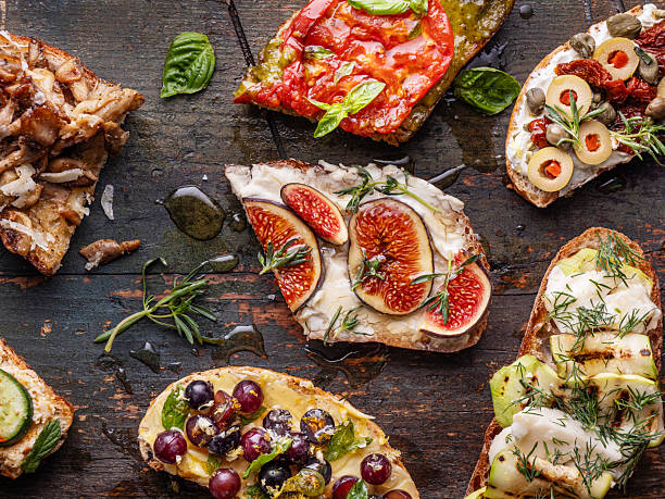 An assortment of gourmet bruschetta and tapas topped with individual ingredients such as fresh figs, sautéed mushrooms, zucchini slices, black olives, green olives, red grapes, roasted red peppers, cream cheese, white cheese, sun-dried tomatoes, capers and fresh herbs, all arranged on a rustic dark wood background. Colorful Mediterranean appetizer selection, overhead view, artisanal food styling, no people.