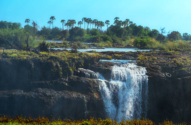 A dramatic view of a cascading waterfall flowing over dark rocks with green riverbank and distant palms under a clear blue sky; a powerful natural scene perfect for travel, nature, and adventure themes.