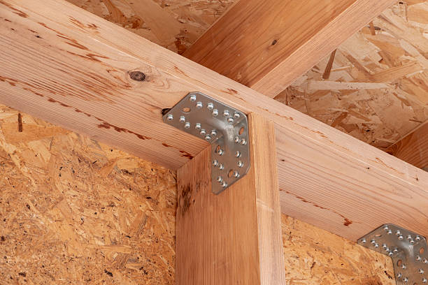 A detailed close-up shot of an interior construction joint showing new wooden beams secured by a heavy-duty, perforated metal angle bracket. The background and wall surface are covered with rough Oriented Strand Board (OSB) sheathing. This image is excellent for illustrating themes related to house construction, carpentry, framing, DIY projects, architecture, structural support, and the proper use of building materials and hardware in home building or renovation.
