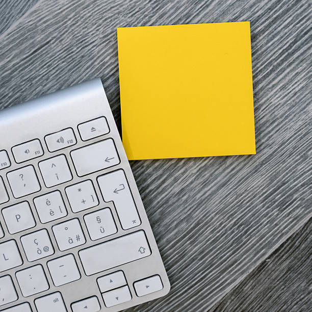 Blank Yellow Sticky Note ready for text mockup next to a silver wireless keyboard on a dark wood desk