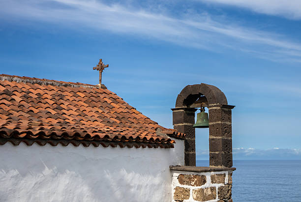 Small old church with white facade, cross and orange roof tiles. Felfry tower with one bell. Stone details. Atlantic ocean and blue sky with light white clouds. San Juan de la Rambla, Tenerife, Spain.