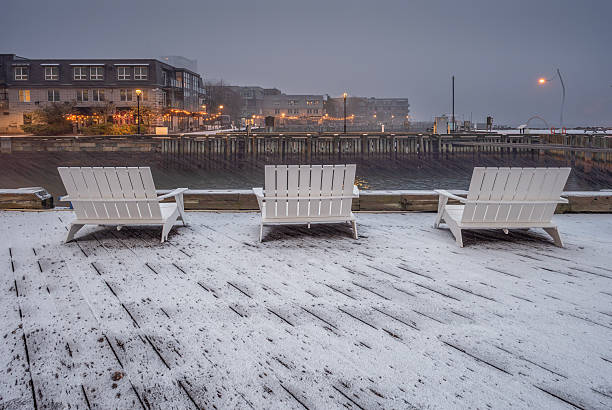 A tranquil winter scene along a lakeside pier where blue street lamps line the walkway. Snow patches dot the pavement as a distant, misty shoreline fades into the overcast horizon.