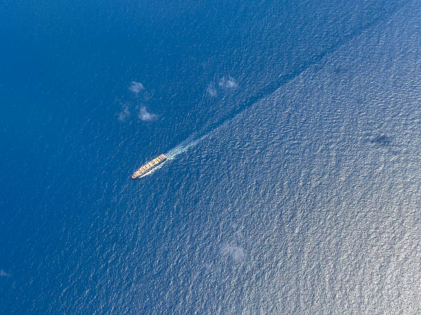 A striking top-down (bird's-eye) aerial perspective of a massive container ship cutting through the deep blue waters of the open ocean. The large vessel leaves a prominent white wake trailing behind it, symbolizing movement and speed. The image perfectly illustrates the scale of global commerce, international trade, maritime shipping, and the complexity of the worldwide supply chain. This minimalist shot is ideal for concepts related to shipping, logistics, technology, and ocean travel.