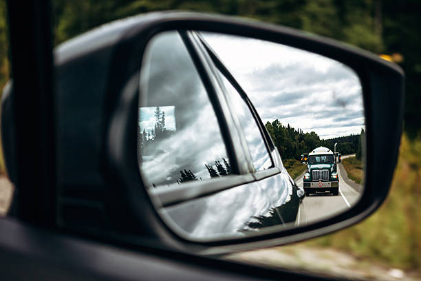A truck's side mirror is reflecting the road ahead. The road is empty and the trees are in the background