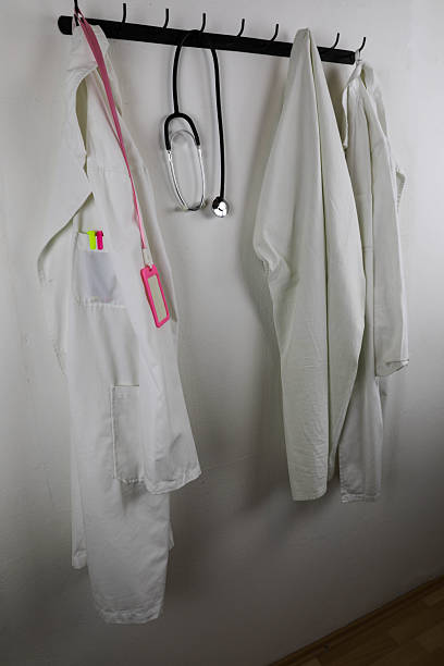 Two White Medical Lab Coats Hanging On A Wall Rack, With A Stethoscope And ID Badge, Representing Healthcare Professionals And Clinical Work Environment.