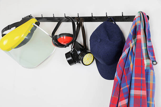 A neatly arranged set of personal protective equipment hanging on a wall-mounted rack, including a yellow face shield, earmuffs, a respirator mask, a blue cap, and a colorful plaid shirt.