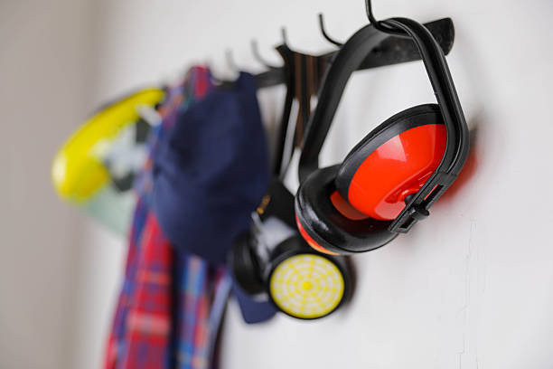 A neatly arranged set of personal protective equipment hanging on a wall-mounted rack, including a yellow face shield, earmuffs, a respirator mask, a blue cap, and a colorful plaid shirt.