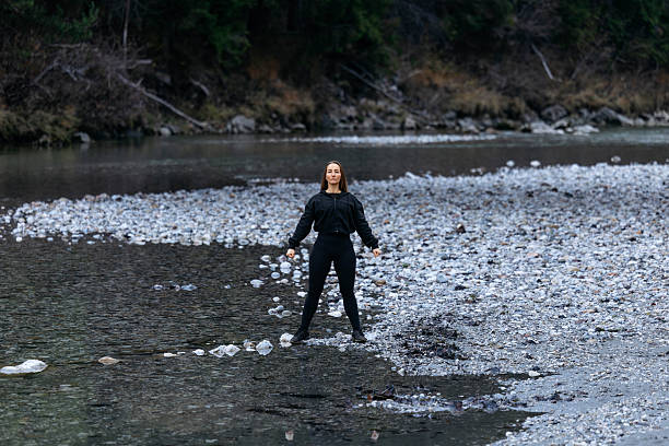 Confident female figure stands in a calm riverbed, surrounded by smooth stones and vibrant greenery, showcasing a harmonious connection with the natural world