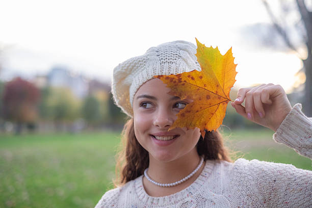 Young woman smiling, holding an autumn leaf over her eye in a park