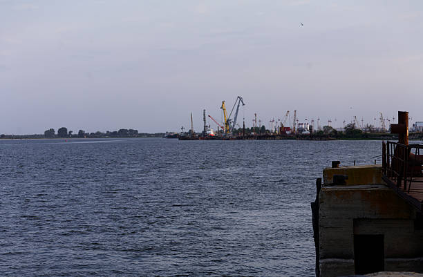 istock Industrial Port Operations: Cargo Ships and Cranes Along Calm River Pier with Overcast Sky 2251435084