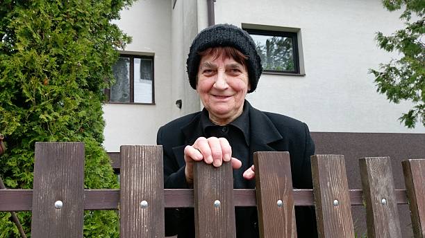 A smiling older woman. Elderly woman in a black coat and knitted hat smiling warmly while leaning on a wooden fence in front of a modern suburban house, framed by tall green shrubs