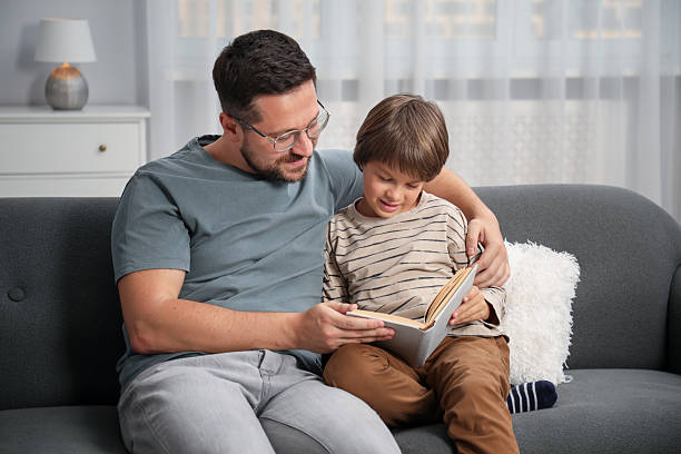 istock Father and son reading book together on sofa at home 2251463739