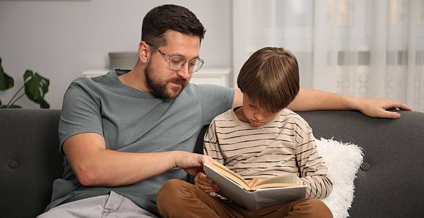 istock Father and son reading book together on sofa at home 2251464334