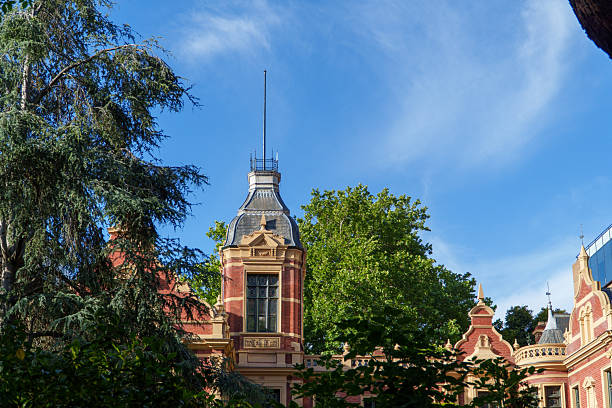 An elaborate red brick building with a decorative octagonal tower and intricate facade, framed by verdant trees against a clear blue sky.