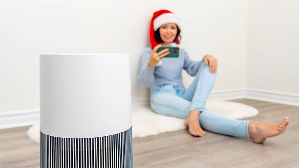 istock Air filter device in modern home setting with a woman wearing a Santa hat, enjoying a smartphone, creating a cozy holiday atmosphere 2251467576