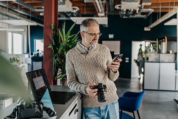 istock Man in sweater looks at phone while holding camera in modern coworking space in Berlin during work hours 2251470604
