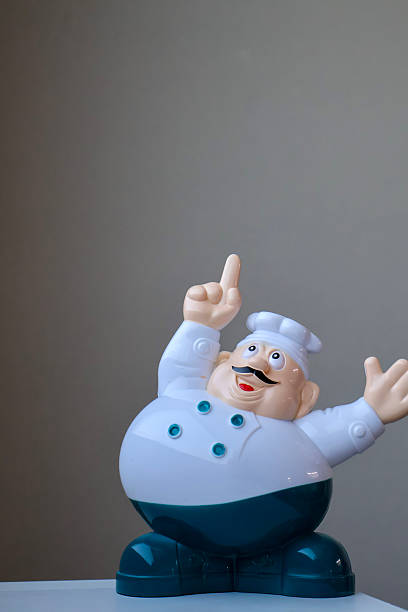 Plastic chef figurine with a happy expression, pointing upwards, isolated on a neutral background. Playful kitchen decoration or cooking-themed toy
