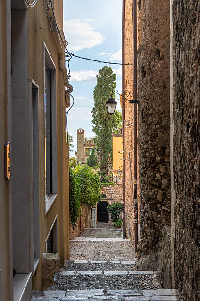 Picturesque narrow alleyway in Taormina, Sicily, featuring steep stone steps lined with colorful ceramic pots, balconies, plants, and charming Mediterranean architecture. The vibrant details and lush greenery create a lively and inviting atmosphere typical of southern Italian hill towns. Bright sunlight and a clear blue sky highlight the beauty of this iconic Sicilian street.