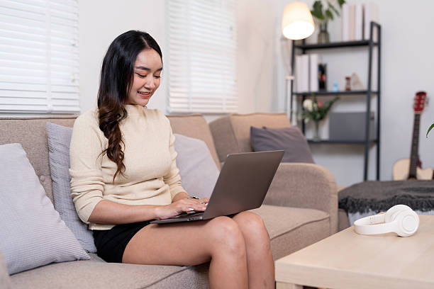 istock Smiling young Asian woman working or browsing on a laptop while relaxing comfortably on a sofa in her modern living room at home. 2251478018