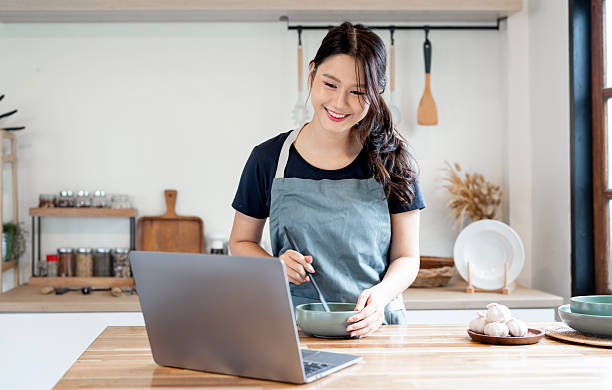 istock Smiling young Asian woman in an apron stirring a meal while simultaneously checking her laptop on the kitchen counter, representing work-life balance and modern cooking. 2251478266