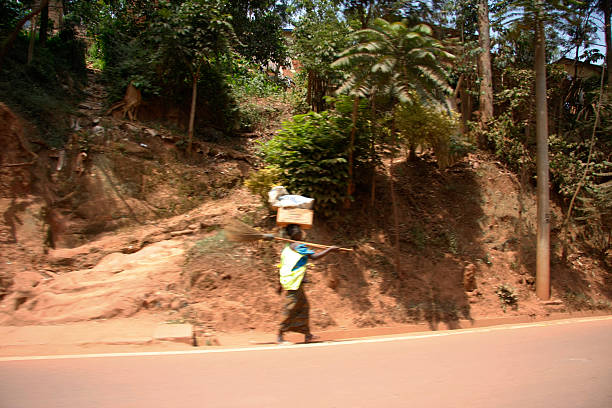 A candid color photograph capturing a woman walking purposefully alongside a paved road, carrying a large box or package balanced on her head. She holds a broom or stick over her shoulder. The scene features a steep red earth embankment and lush tropical foliage, characterizing daily life, traditional methods of transport, and human labor in a rural or suburban setting. The image is a study in perseverance and the pace of life contrasting with the motorized traffic of the nearby road.