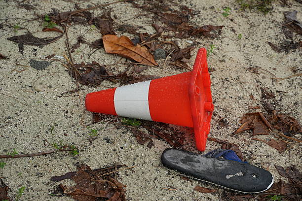 A discarded traffic cone and shoe lie on sandy ground covered with leaves, evoking a sense of neglect and desolation.