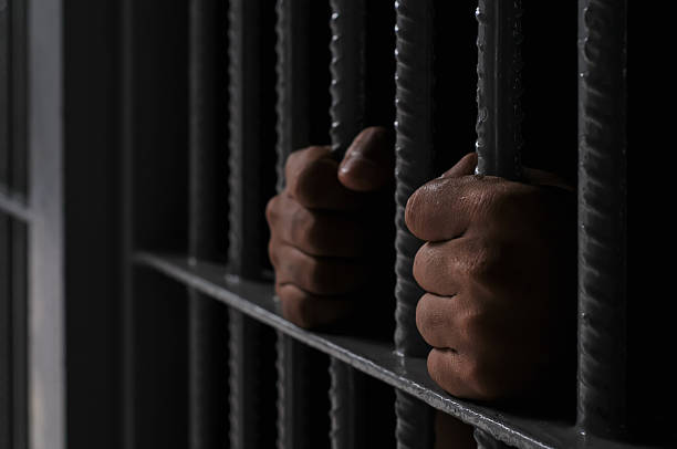 istock prisoner's hands behind bars with black background 2251487864