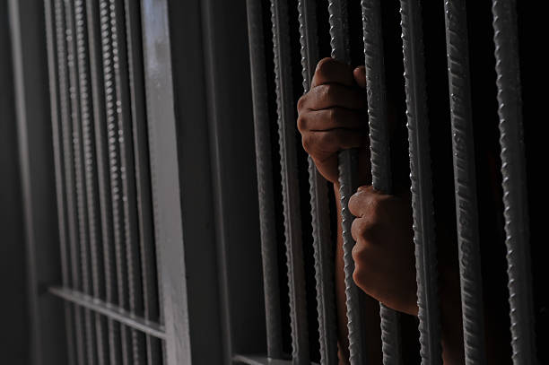 istock prisoner's hands behind bars with black background 2251487870