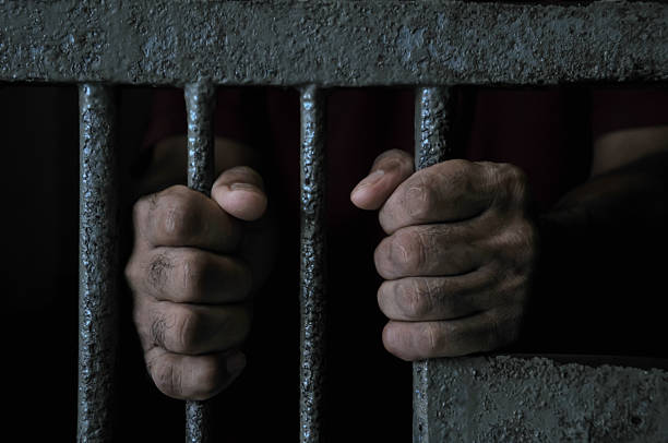 istock prisoner's hands behind bars with black background 2251487879