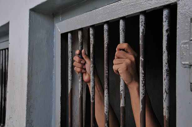 istock prisoner's hands behind bars with black background 2251487897