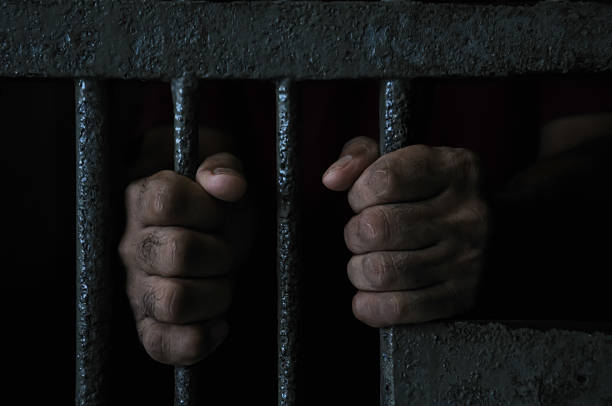istock prisoner's hands behind bars with black background 2251487915