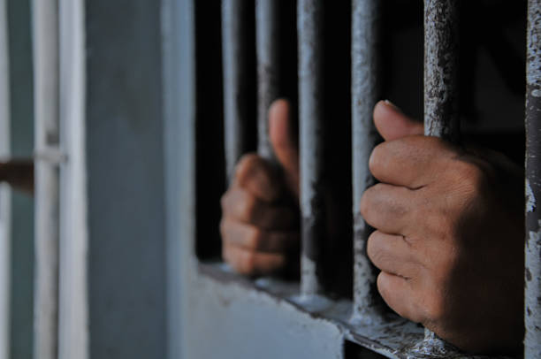 istock prisoner's hands behind bars with black background 2251487959