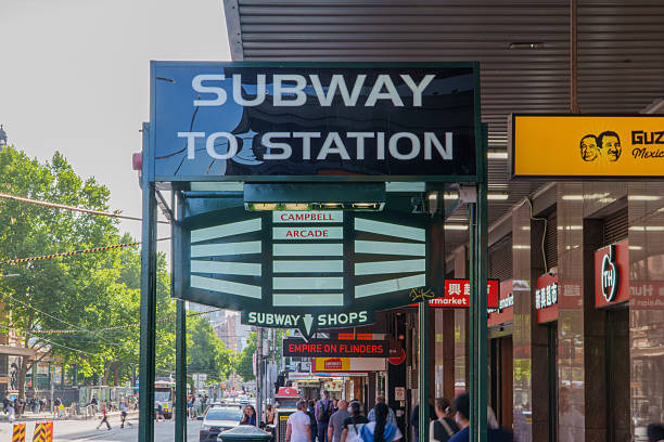 Melbourne, VIC, Australia. December 11, 2025. - An overhead sign for Campbell Arcade and a subway station directs pedestrians on a bustling, sunlit city street with shops.